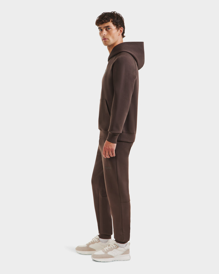 A man stands sideways wearing the Rudsak BRIAR - 9525001 Espresso M unisex hoodie and matching joggers in a premium cotton blend, styled with light beige sneakers against a plain white background for a relaxed, casual look.