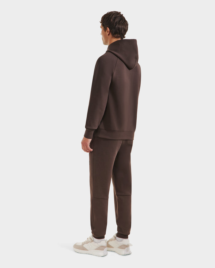A person faces away wearing the Rudsak BRIAR - 9525001 Espresso M unisex hoodie and jogger set, paired with white and beige sneakers. The plain white background highlights the relaxed, casual vibe.