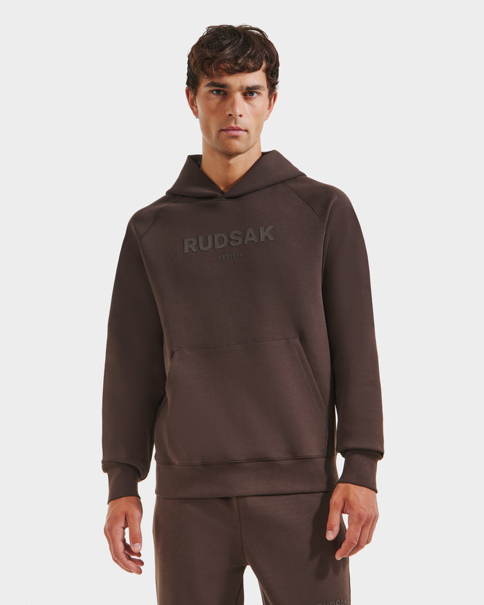 A man faces forward in the Rudsak BRIAR - 9525001 Espresso M unisex hoodie and matching pants. “RUDSAK” is printed on the premium cotton-blend sweatshirt. The background is plain white, creating an elevated casual vibe.