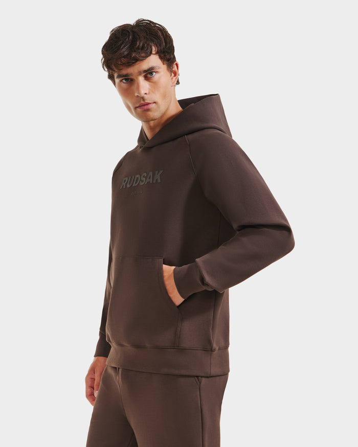 A young man stands against a plain white background wearing the Rudsak BRIAR - 9525001 Espresso M unisex hoodie, crafted from premium cotton blend, featuring RUDSAK on the front, looking to the side with one hand in his pocket for a casual vibe.