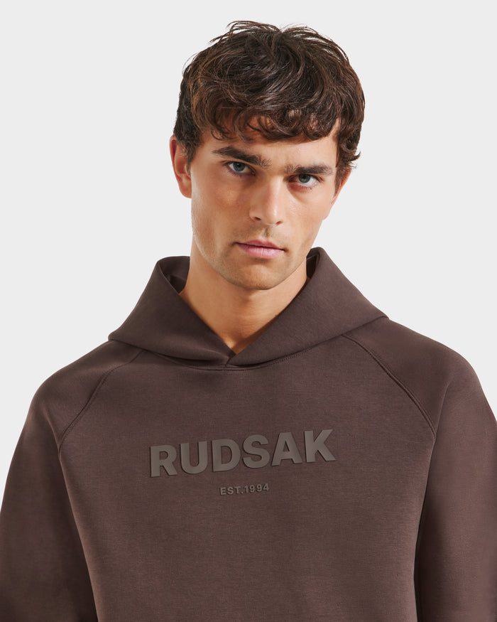 A young man with short brown hair models the Rudsak BRIAR - 9525001 Espresso M unisex hoodie, made from premium cotton blend with raised RUDSAK and EST. 1994 lettering on the chest, against a plain white background for a casual vibe.