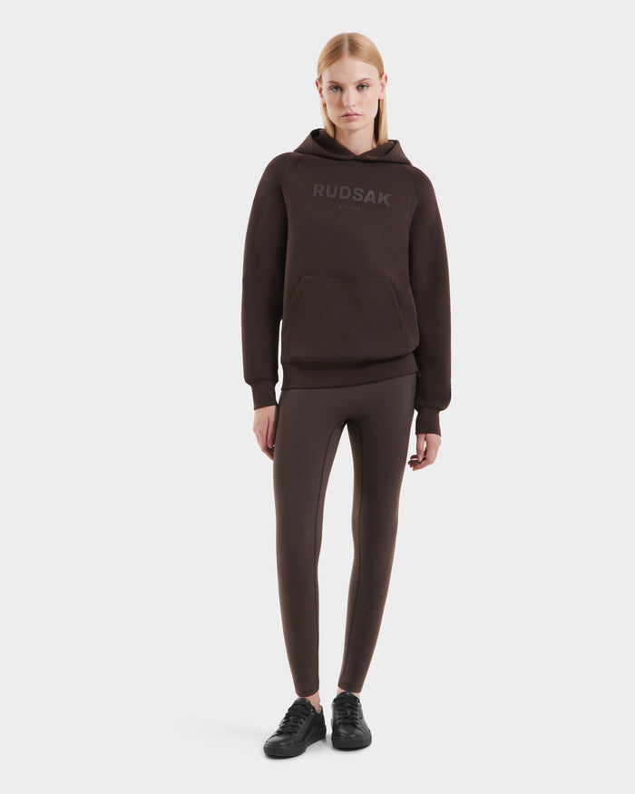 A woman faces forward in the Rudsak BRIAR - 9525001 Espresso W premium cotton blend unisex hoodie, styled with matching brown leggings and black sneakers, set against a plain white background.