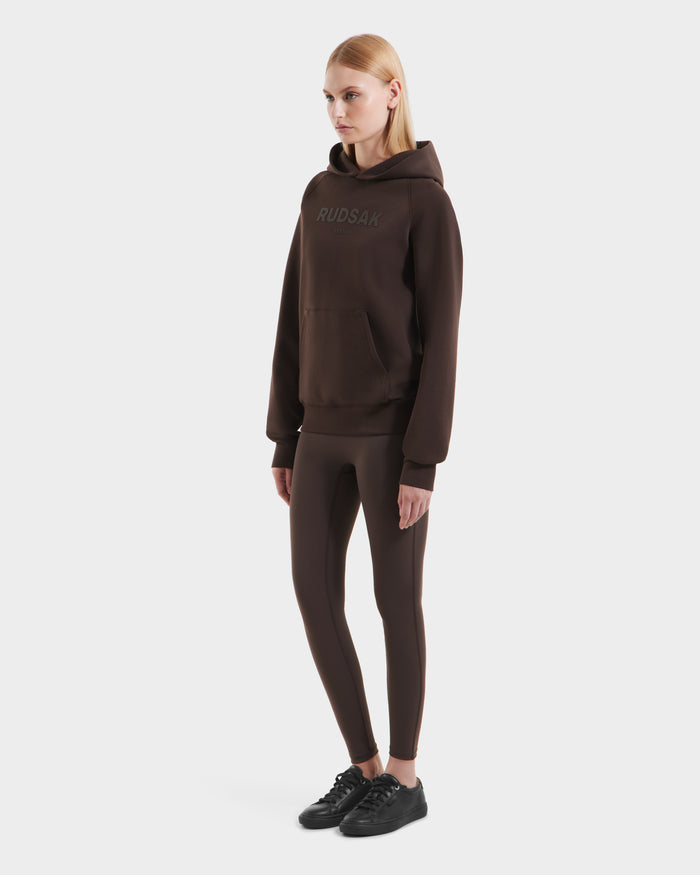 A woman stands against a plain white background wearing the Rudsak BRIAR - 9525001 Espresso W premium cotton blend hoodie, matching brown leggings, and black sneakers.
