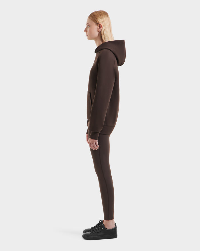 A person with straight blonde hair stands in profile, wearing the Rudsak BRIAR - 9525001 Espresso W unisex hoodie and matching leggings, paired with black sneakers, all set against a plain white background.
