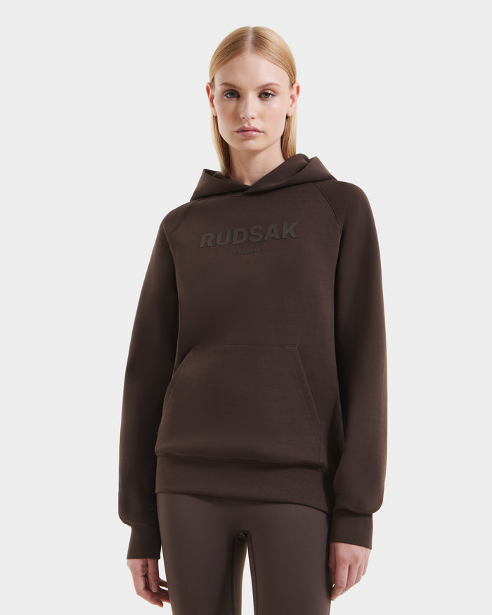 A woman with straight blonde hair wears the Rudsak BRIAR - 9525001 Espresso W hoodie and matching pants, crafted from a premium cotton blend, standing against a plain white background.