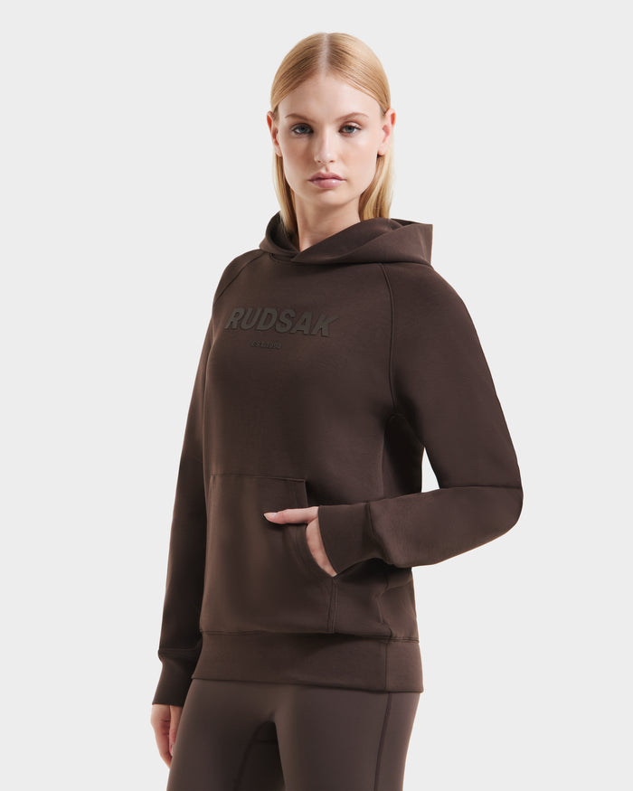 A woman models the Rudsak BRIAR - 9525001 Espresso W unisex hoodie in premium cotton blend with RUDSAK front print, standing against a white background, straight blonde hair, one hand in her pocket.