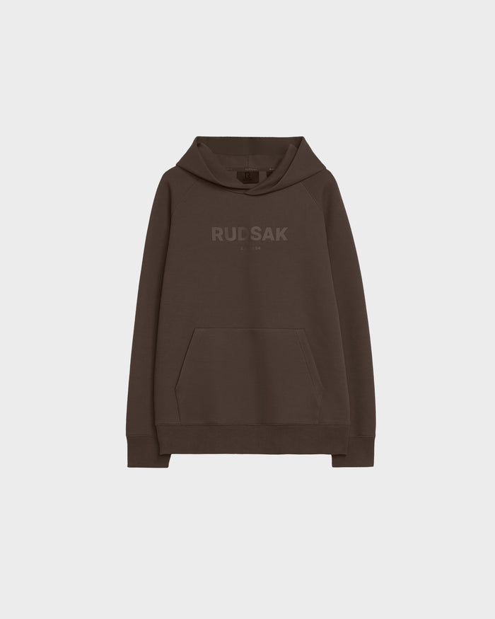 The Rudsak BRIAR - 9525001 Espresso W is a dark brown unisex hoodie with a front kangaroo pocket and RUDSAK in matching brown on the chest, shown on a plain light gray background.