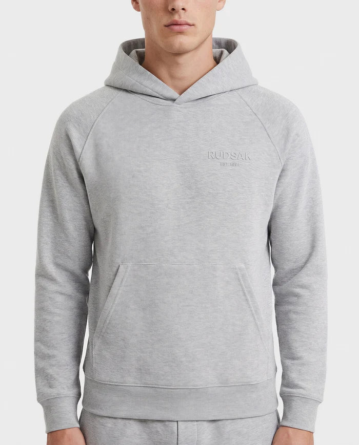 A person wears the Rudsak BRIAR - 9525001 hoodie in heather grey (M), a unisex premium cotton blend with a front pocket and Rudsak embroidered on the left chest, standing against a white background for a casual look.
