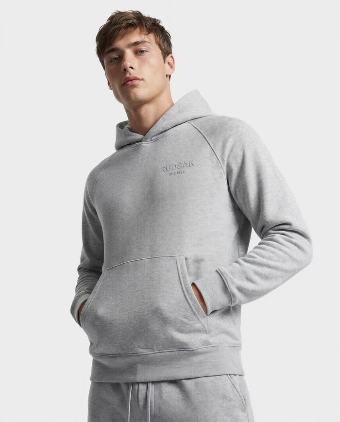 A young man wears the Rudsak BRIAR - 9525001 hoodie in heather grey (M), made from a premium cotton blend, with hands in the front pocket. "RUDSAK EST. 1994" is subtly printed on the chest against a plain white background.