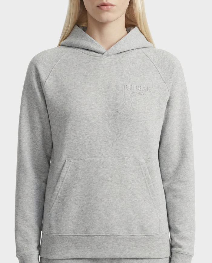 A woman models the Rudsak BRIAR - 9525001 Heather Grey W unisex hoodie, crafted from a premium cotton blend with a front pocket and RUDSAK embroidery on the chest, set against a plain white background.