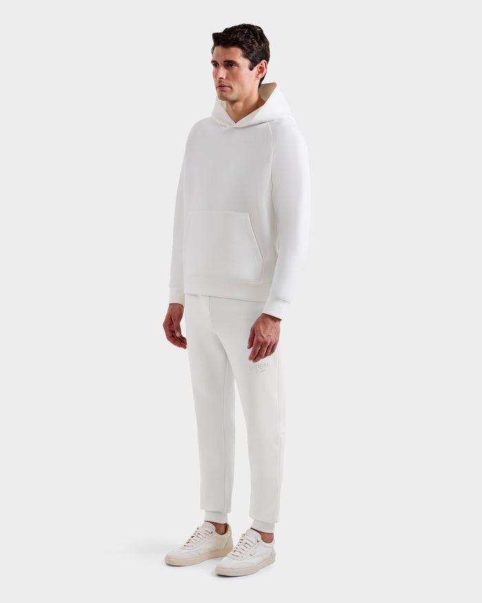 A man models the Rudsak BRIAR - 9525001 Iceland M: an all-white unisex premium cotton blend hoodie and jogger pants, paired with white sneakers. He stands against a plain white background, looking slightly to the side.