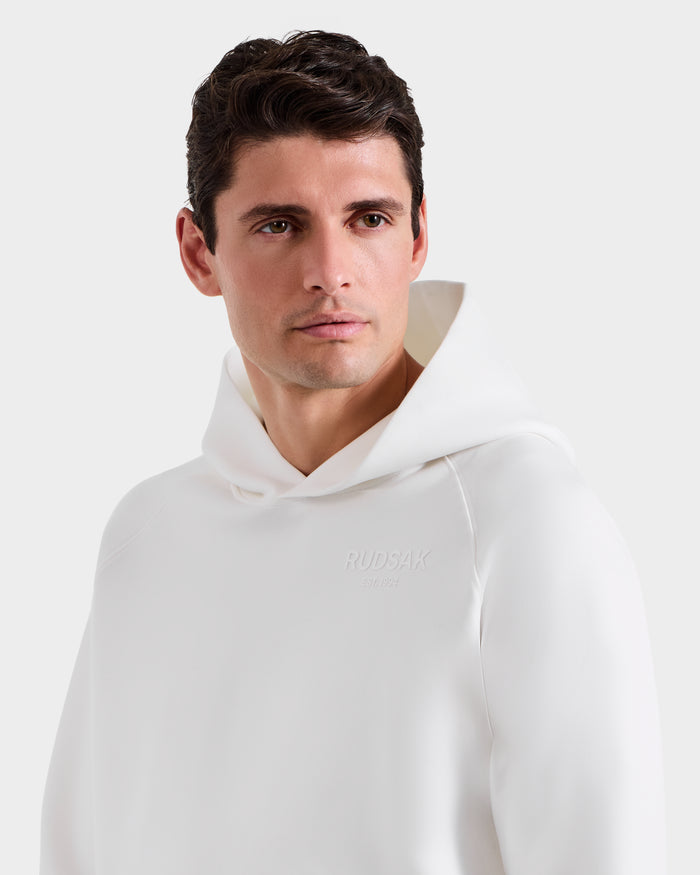 A man models the Rudsak BRIAR - 9525001 Iceland M unisex white hoodie with embroidered branding, glancing to the side against a plain white background for a relaxed, casual style.