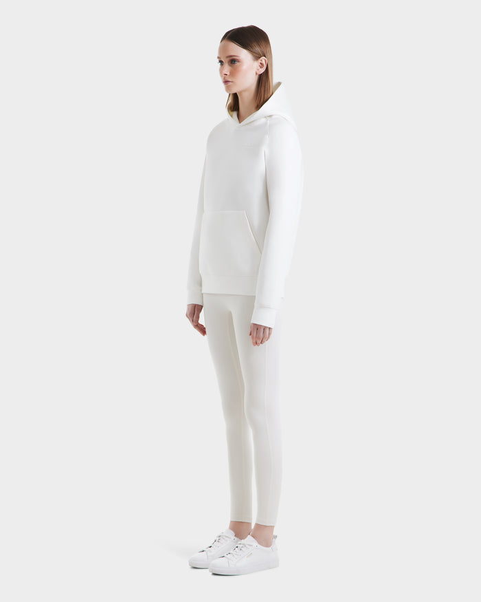 A woman stands against a plain white background wearing the Rudsak BRIAR - 9525001 Iceland W hoodie, fitted pants, and white sneakers, facing slightly right with hands by her sides for a fresh, casual look.