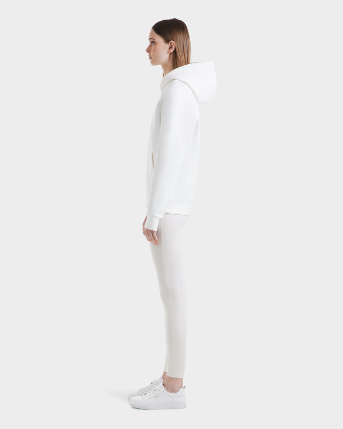 A woman in profile faces left, wearing the Rudsak BRIAR - 9525001 Iceland W all-white unisex hoodie and fitted pants, paired with white sneakers, set against a plain light gray background for a modern casual look.