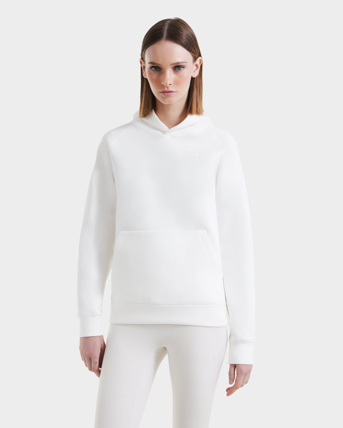 A woman faces forward, wearing a Rudsak BRIAR - 9525001 Iceland W white unisex hoodie made from premium cotton blend with a front pocket and matching white leggings, set against a plain light gray background.