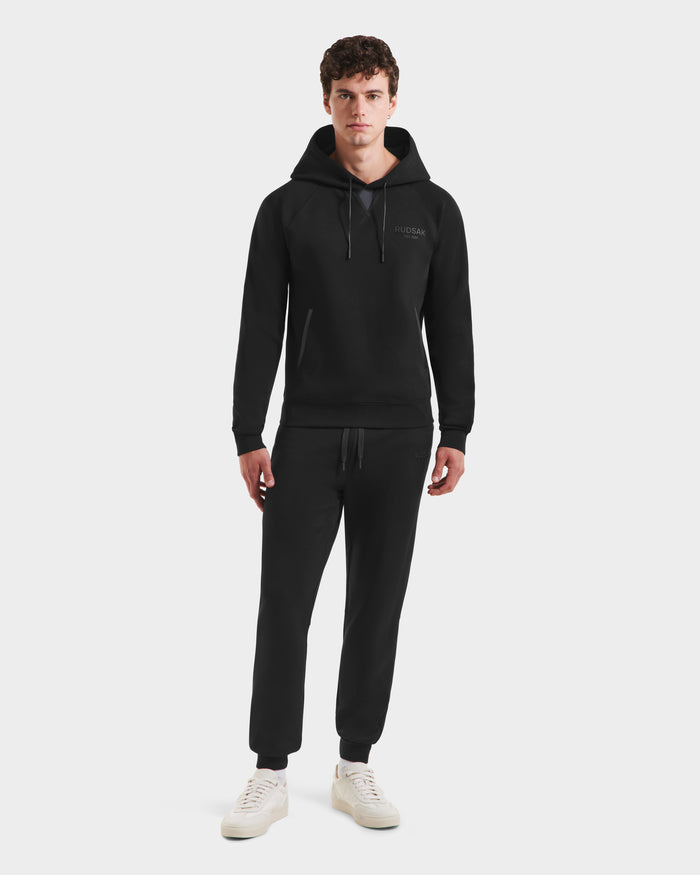 A young man faces forward against a white background, wearing the Rudsak BROOKLYN - 6525593 Black hoodie and matching sweatpants with white sneakers.