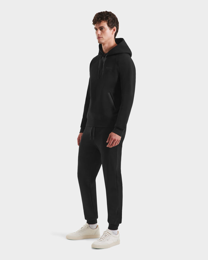A man stands against a plain white background wearing the Rudsak BROOKLYN - 6525593 Black hoodie and matching jogger pants, styled with white sneakers.