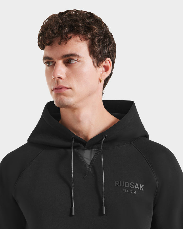 A young man with short curly brown hair wears the Rudsak BROOKLYN - 6525593 Black hoodie, featuring bold chest branding, as he looks to his left against a plain white background.