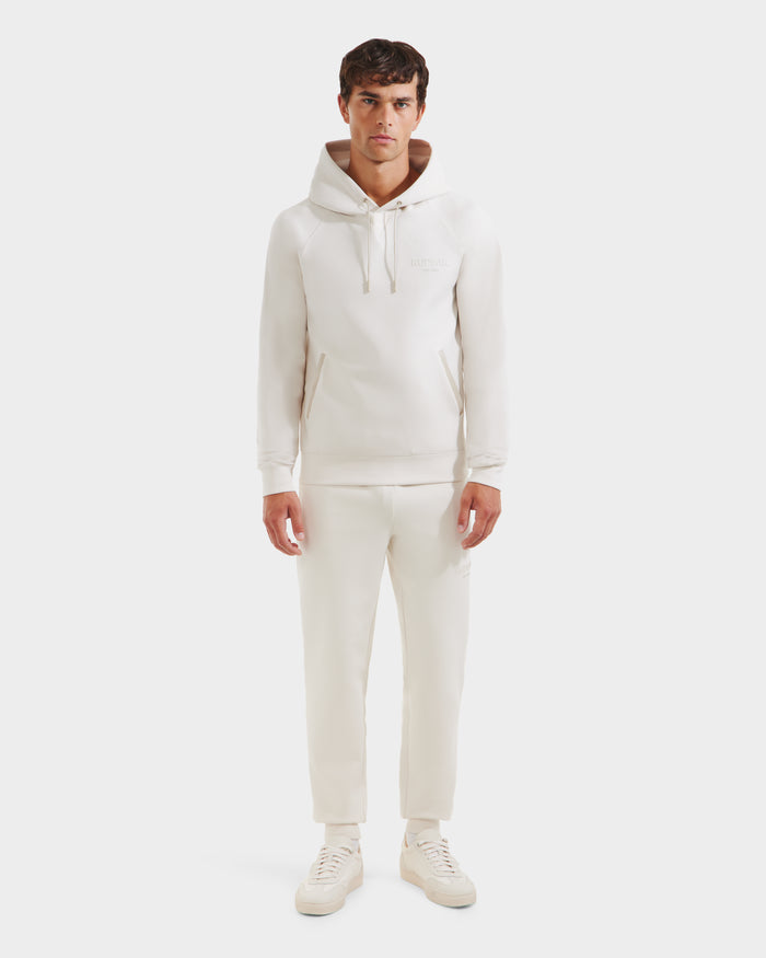 A man faces forward against a plain white background, wearing Rudsak’s BROOKLYN - 6525593 Vanilla hoodie and matching sweatpants, paired with white sneakers.