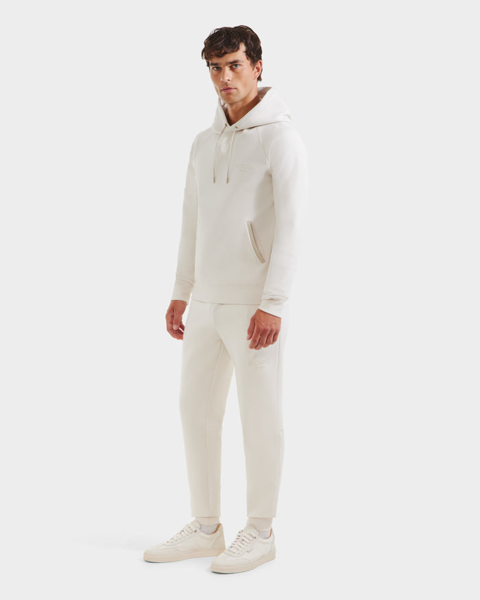 A man models the Rudsak BROOKLYN - 6525593 Vanilla hoodie and jogger set, paired with cream sneakers, facing slightly to the side with a neutral look against a plain white background.