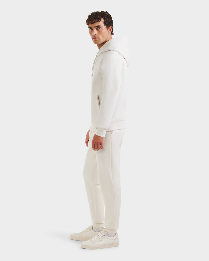 A man wearing Rudsaks BROOKLYN - 6525593 Vanilla set stands sideways in matching off-white hoodie and sweatpants with white sneakers, facing the camera against a plain white background.