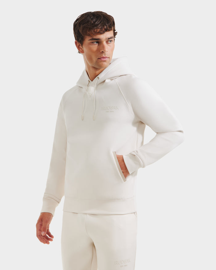 A man models the Rudsak BROOKLYN - 6525593 Vanilla hoodie and sweatpants set, standing against a white background with one hand in his pocket, embodying a clean urban streetwear look as he gazes to the side.