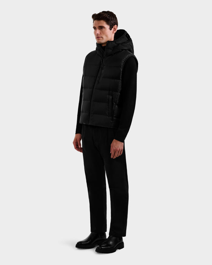 A man stands slightly angled, wearing the Rudsak BROOKS - M125520 Black hooded down vest over a black long-sleeve shirt, black pants, and black shoes, set against a plain light gray background.