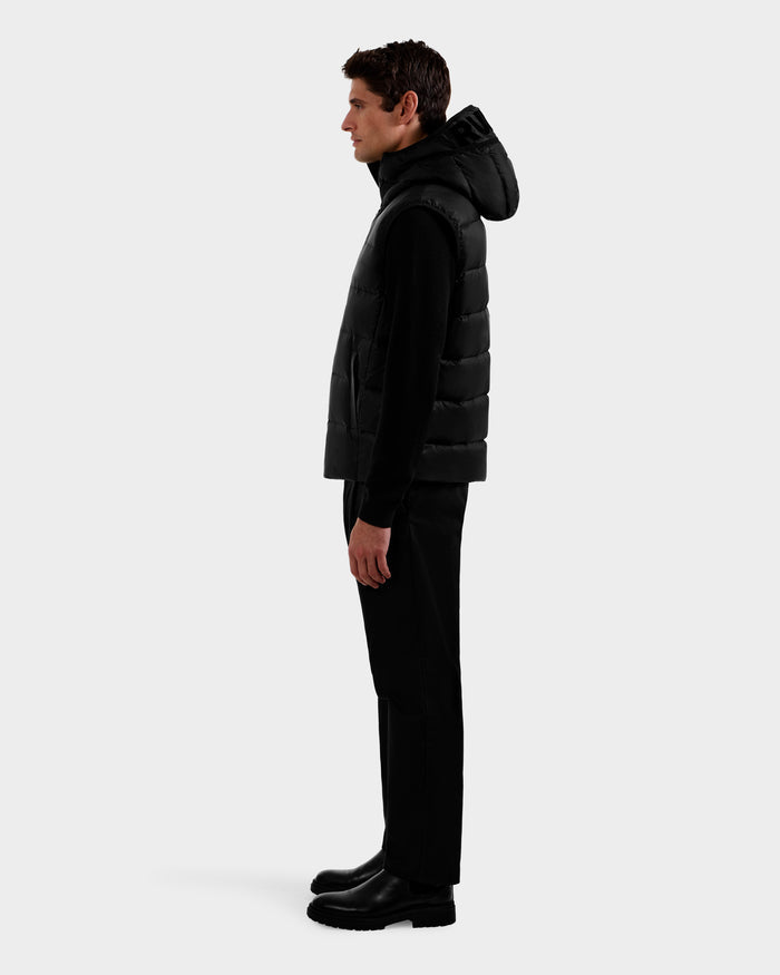 A man stands in profile against a plain background, wearing the Rudsak BROOKS - M125520 Black men’s down puffer vest with a hood made from recycled nylon, paired with black pants and black boots.