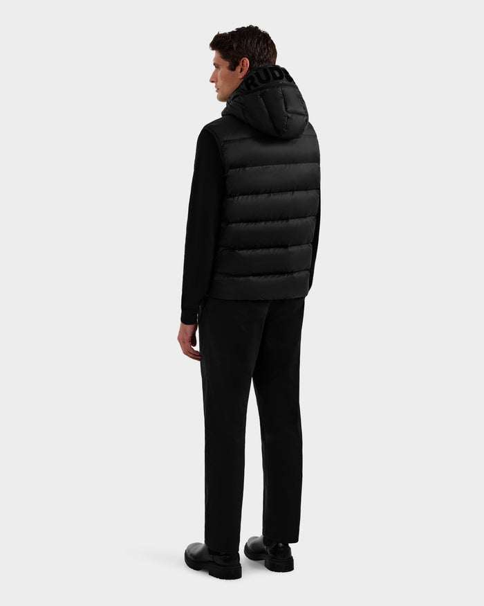A man faces away, wearing the Rudsak BROOKS - M125520 Black hooded down vest made from recycled nylon, paired with a black long-sleeve shirt, pants, and shoes against a plain light gray background.