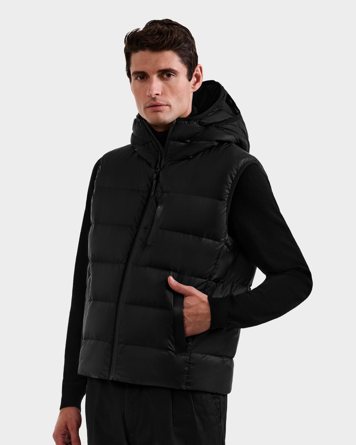 A man wearing the Rudsak BROOKS - M125520 Black down puffer vest over a black long-sleeve shirt stands with one hand in his pocket, looking at the camera against a plain light background.