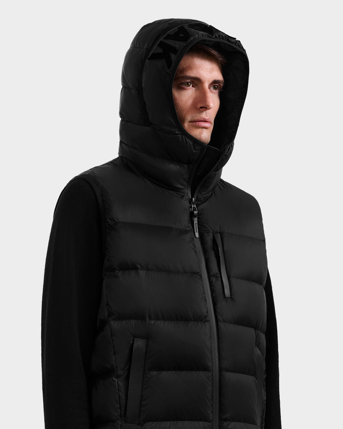 Wearing the Rudsak BROOKS - M125520 Black hooded down vest made from recycled nylon with a front zipper, a person looks to the side against a plain light background.