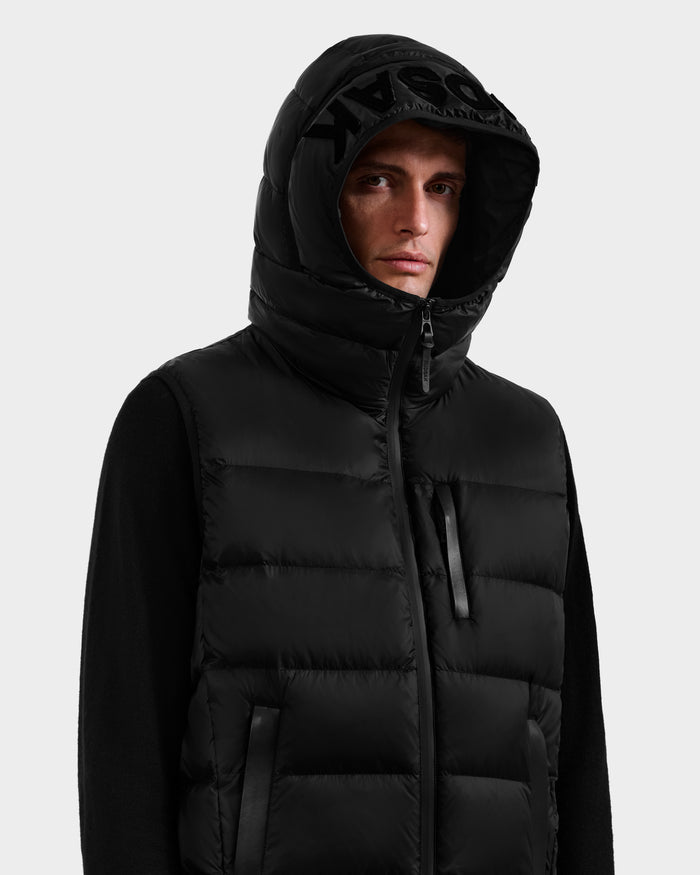 A man wearing the Rudsak BROOKS - M125520 Black men’s hooded down puffer vest, made from recycled nylon, with a high collar, front zipper, and subtle front logo; hood up against a plain light background.