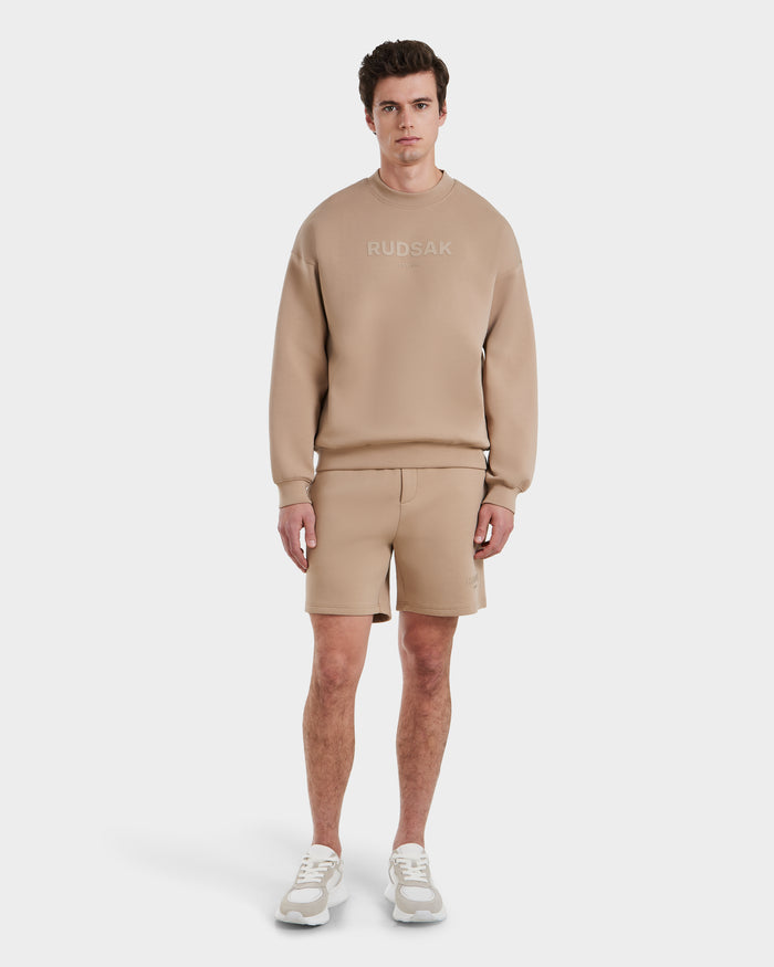 A man stands against a white background wearing the BRYN - 9524007 Almond M crewneck sweatshirt by Rudsak, paired with matching beige shorts and white sneakers—an everyday, genderless look.