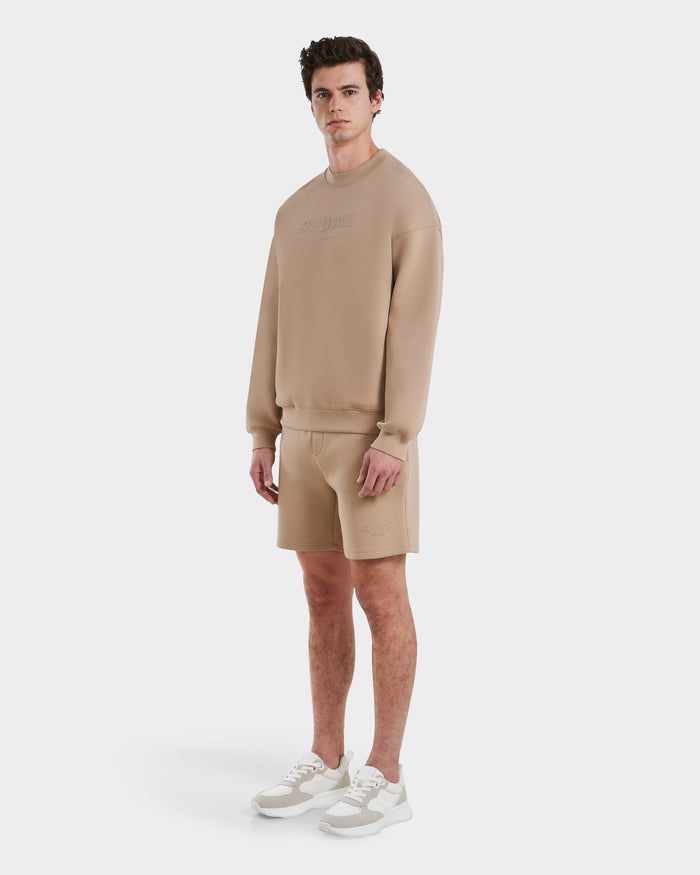 A man models the Rudsak BRYN - 9524007 Almond M set—beige cotton crewneck sweatshirt and shorts—paired with white/beige sneakers. He faces the camera neutrally. Genderless sizing offers all-day comfort.