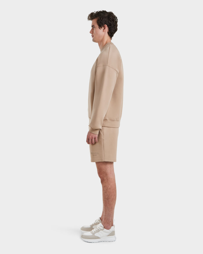 A man in profile against a white background wears the Rudsak BRYN - 9524007 Almond M crewneck sweatshirt with genderless sizing, matching shorts, and beige and white sneakers.