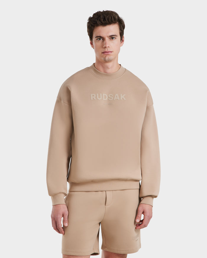 A young man stands against a plain white background, wearing the Rudsak BRYN - 9524007 Almond M cotton crewneck sweatshirt and matching beige shorts, effortlessly showcasing a stylish everyday look.
