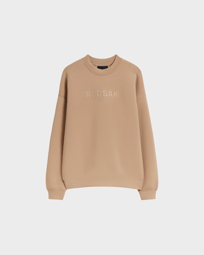 The Rudsak BRYN - 9524007 Almond M is a beige cotton crewneck sweatshirt with long sleeves, ribbed cuffs and hem, and tonal “RUDSAK” chest embroidery. Genderless fit for everyday comfort, shown here against a white background.