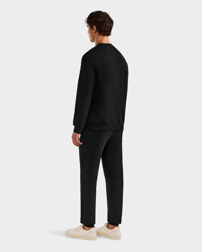 A person faces away from the camera, wearing the Rudsak BRYN - 9524007 Black M crewneck sweatshirt with matching black pants and white sneakers, set against a plain light gray background.