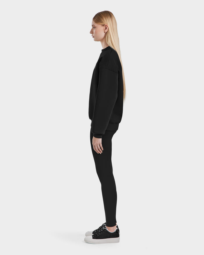 A woman with long blonde hair stands in profile against a plain white background, wearing the Rudsak BRYN - 9524007 Black W sweatshirt, paired with black leggings and black sneakers with white soles for an effortless everyday look.