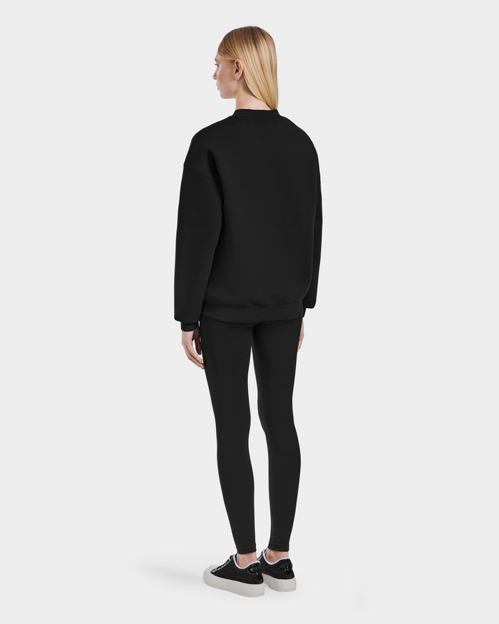 A person with straight, blonde hair wears the Rudsak BRYN - 9524007 Black W crewneck sweatshirt, black leggings, and black-and-white sneakers—modeling a genderless everyday look against a plain white background.