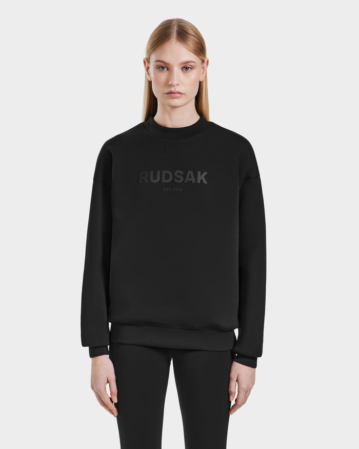 A woman with straight blonde hair wears the Rudsak BRYN - 9524007 Black W cotton crewneck sweatshirt with RUDSAK EST. 1994 on the front, paired with black pants, standing against a plain white background.
