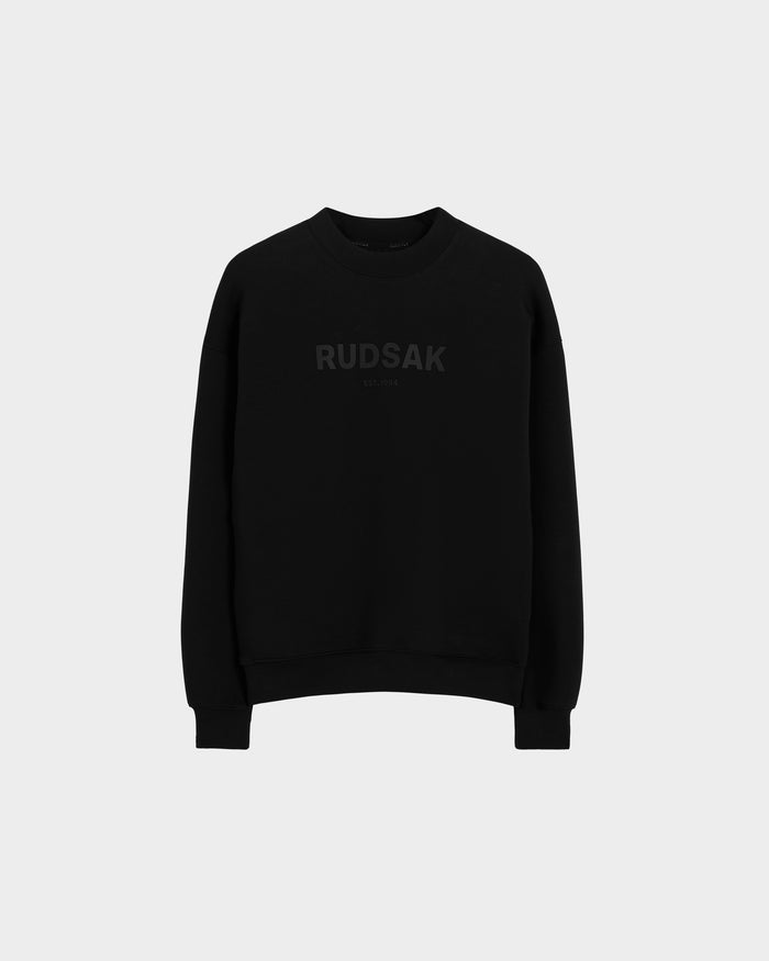 The BRYN - 9524007 Black W crewneck sweatshirt by Rudsak features subtle dark RUDSAK and EST. 1994 prints on the front, crafted from black cotton with genderless sizing—displayed on a light grey background for versatile everyday style.