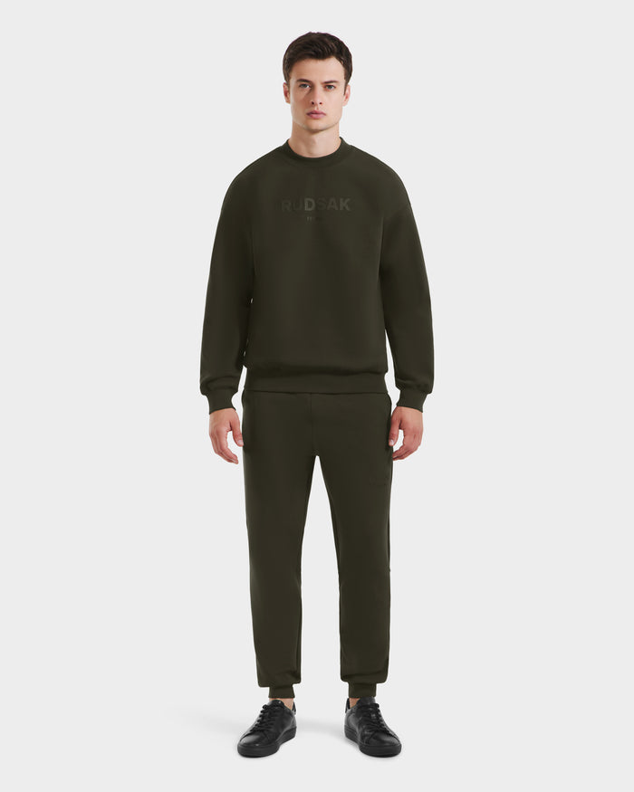 A man faces forward against a plain white background, wearing Rudsaks BRYN - 9524007 set in Dark Olive (M)—a matching cotton crewneck sweatshirt and joggers—styled with black sneakers.