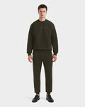 A man faces forward against a plain white background, wearing Rudsaks BRYN - 9524007 set in Dark Olive (M)—a matching cotton crewneck sweatshirt and joggers—styled with black sneakers.