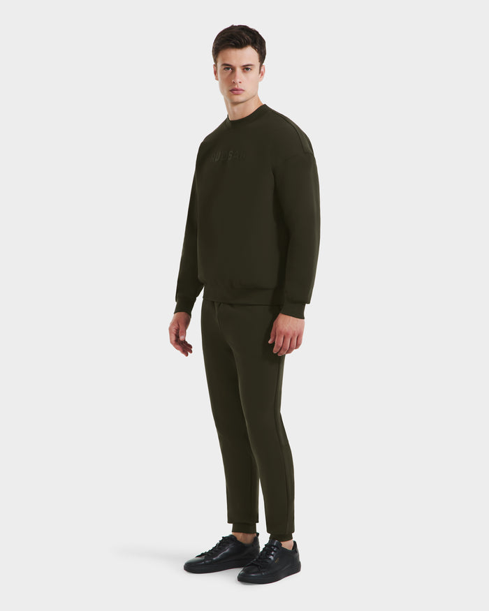 A man wears the Rudsak BRYN - 9524007 Dark Olive M set—an everyday fit, genderless cotton crewneck sweatshirt and pants—with black sneakers, facing slightly to the side against a plain white background.