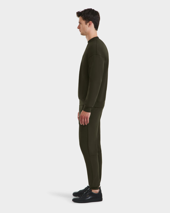 A man stands in profile facing left, wearing the Rudsak BRYN - 9524007 Dark Olive M cotton crewneck sweatshirt and matching joggers with black sneakers, showcasing an everyday fit against a plain white background.