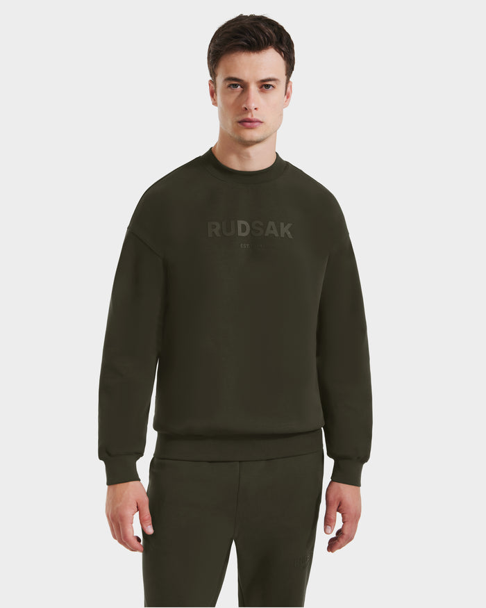 A model faces forward wearing Rudsak's BRYN - 9524007 sweatshirt and matching pants in dark olive. The genderless cotton crewneck features subtle, tonal RUDSAK lettering across the chest against a plain white background.