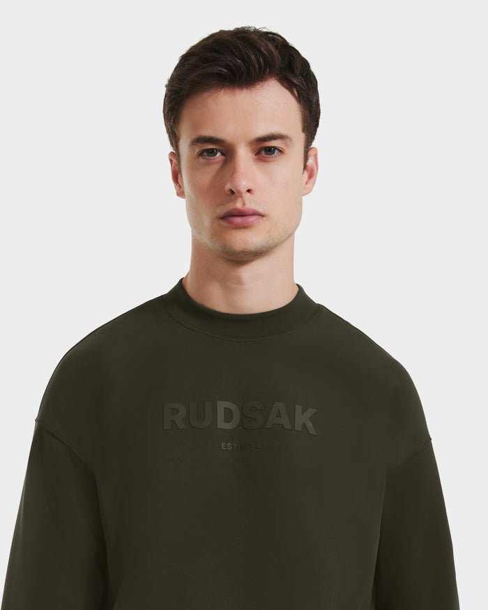 A young man with short brown hair wears the Rudsak BRYN - 9524007 crewneck sweatshirt in Dark Olive (size M), featuring an embossed logo on the front. He faces forward against a plain white background, highlighting its genderless, everyday fit.