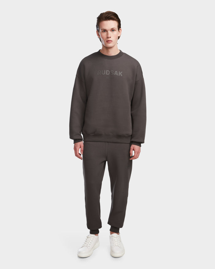 A young man faces forward in a Rudsak BRYN - 9524007 Grey M cotton crewneck sweatshirt, matching joggers, and white sneakers, highlighting timeless style against a plain white background.