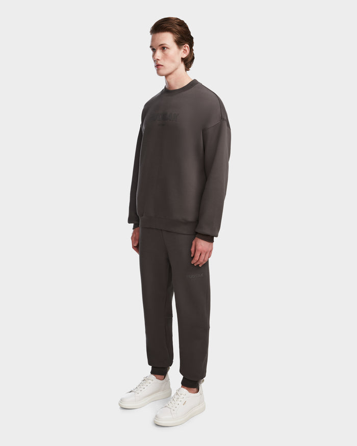 A person stands slightly to the side, wearing the Rudsak BRYN - 9524007 Grey M cotton crewneck sweatshirt and sweatpants set with white sneakers, against a plain white background.
